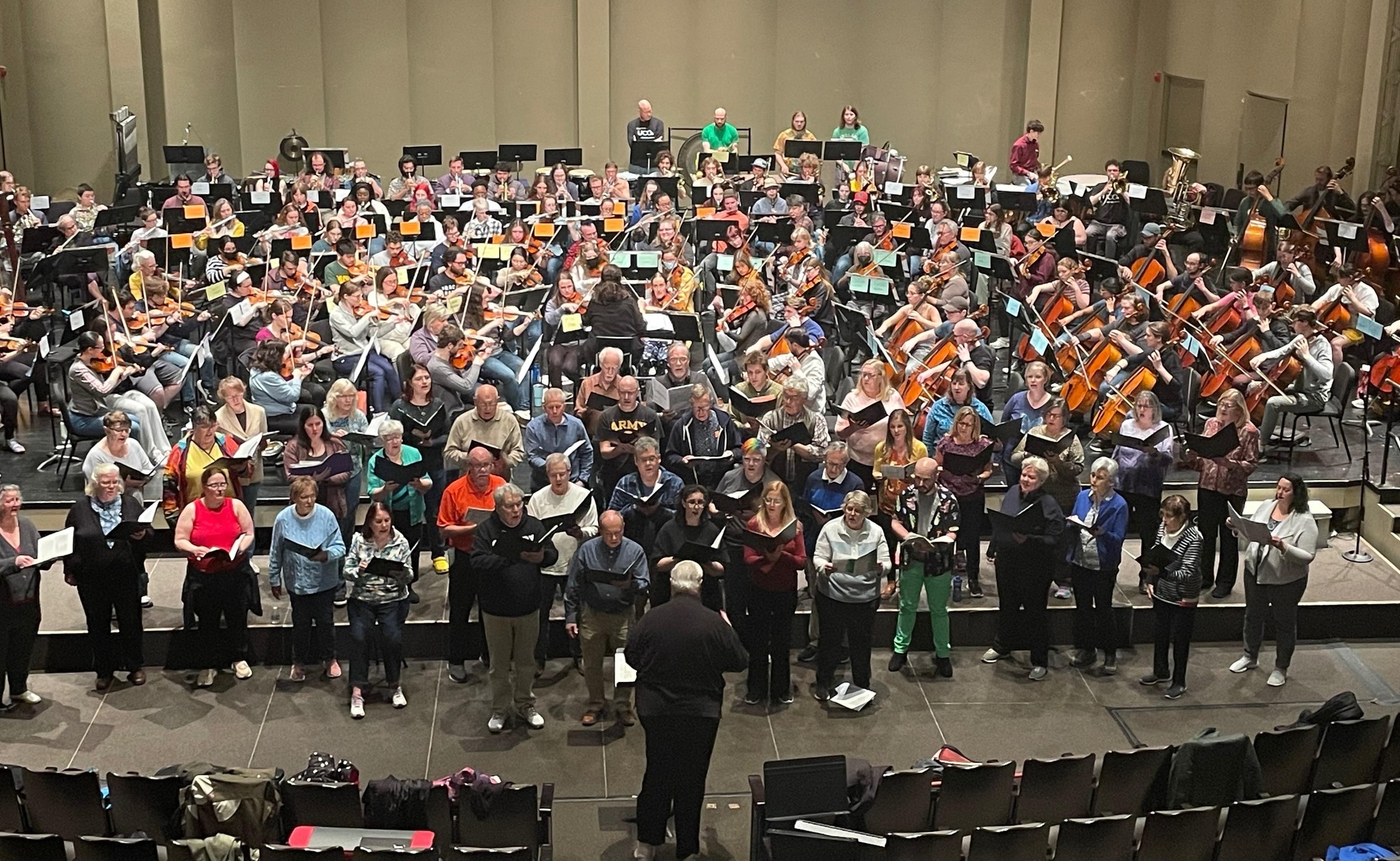 A large orchestra rehearses onstage behind a choir, which stands in front of the stage.